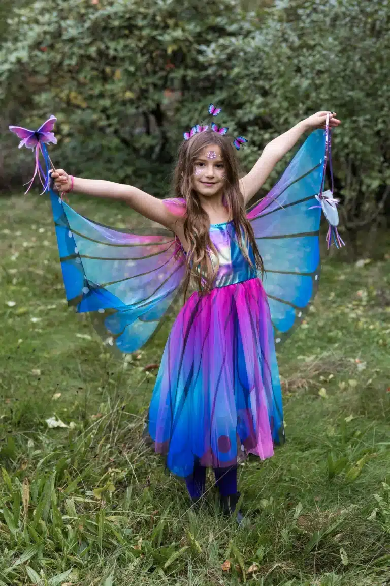 Meisje draagt great pretenders verkleedkleding: een blauw-paarse vlinderjurk met uitgespreide vleugels en vlinderstaf in het bos.