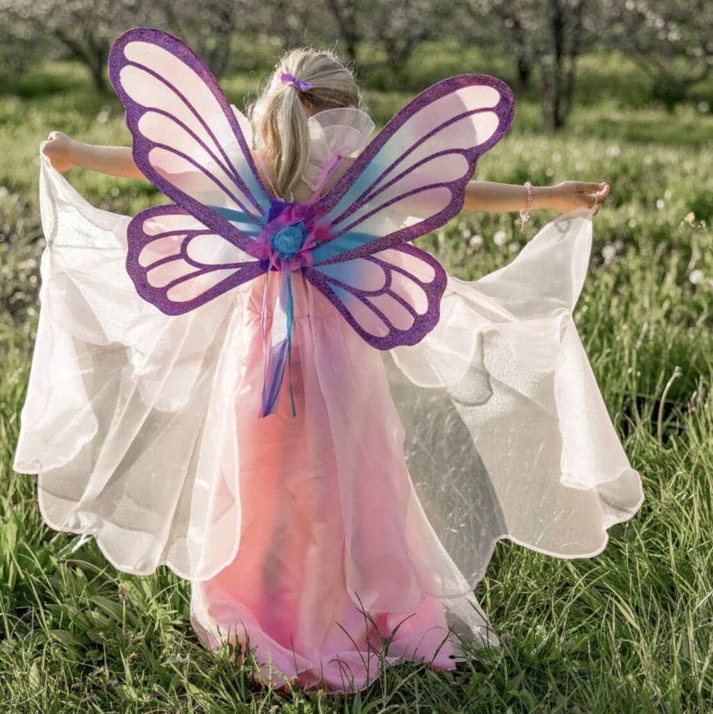 Meisje van achteren in een roze vlinderjurk met paarse vleugels, terwijl ze de geschulpte rok spreidt in het gras