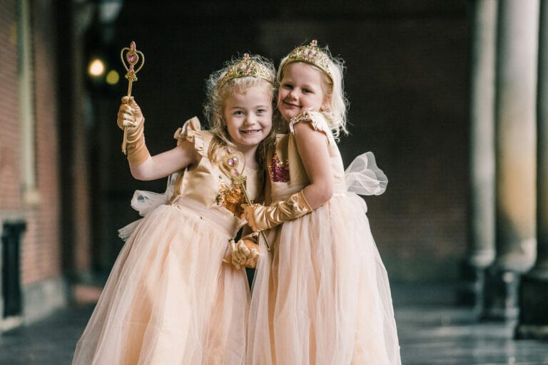 Twee meisjes dragen een gouden princessenjurk van Het Betere Merk met kroon, lange handschoenen en een toverstaf.