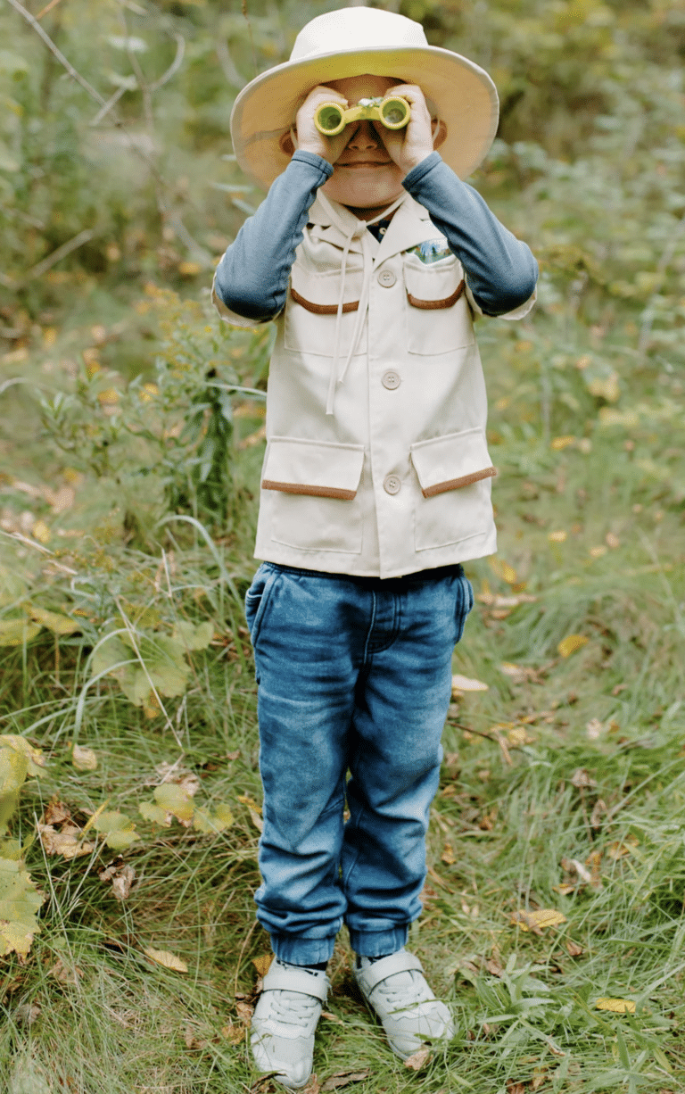Jongen draagt de Forest Guardian verkleedset met safarihoed, beige vest en felgroene verrekijker in het bos.