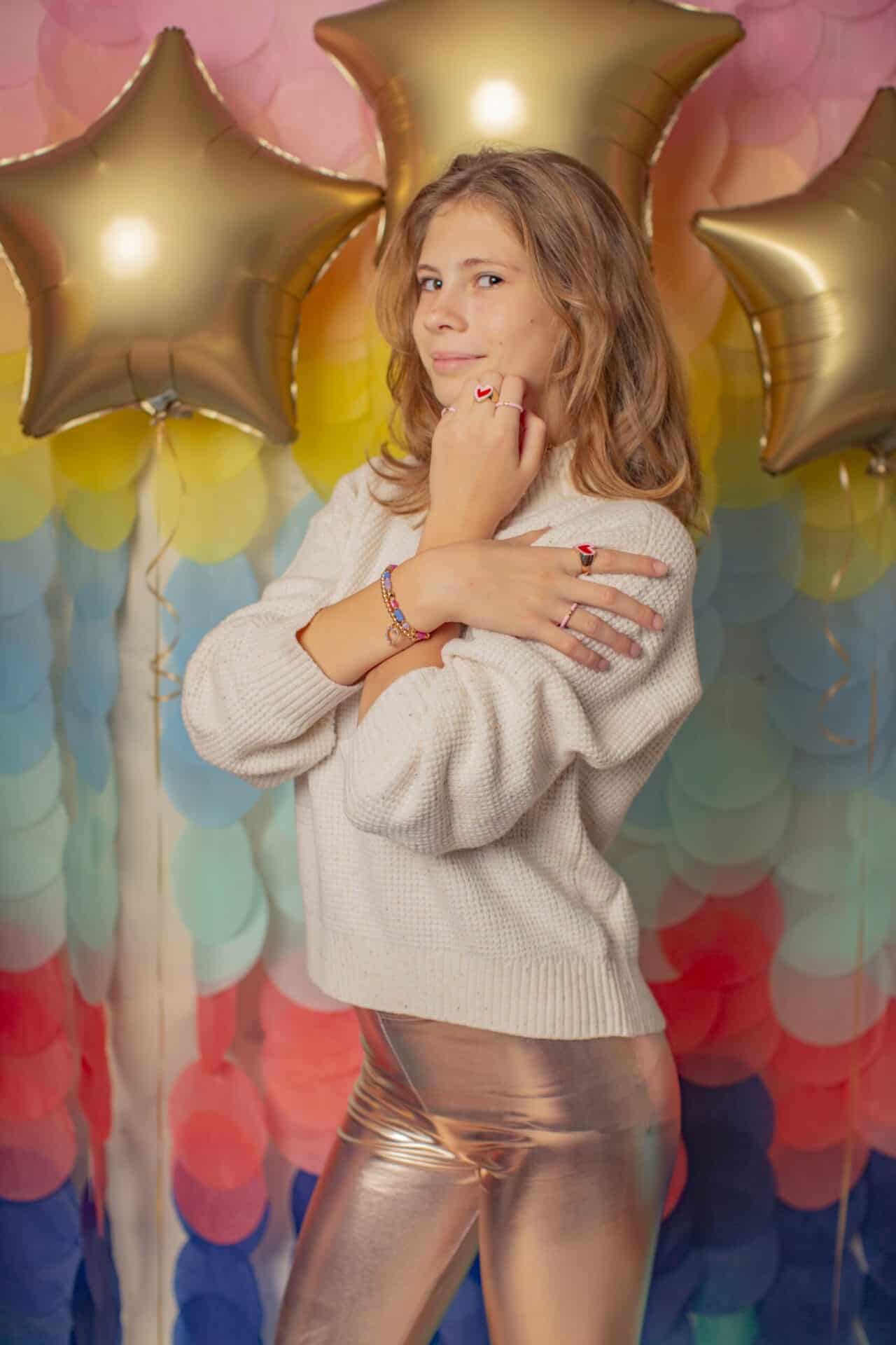 Model toont de Great Pretenders kinderringen met hartje aan haar vingers, tegen een kleurrijke achtergrond met gouden sterrenballonnen.