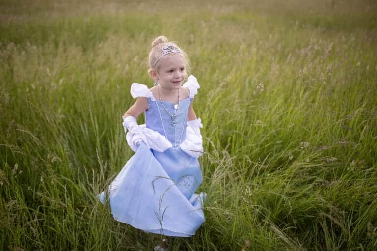 Meisje draagt een blauwe cinderella jurk van Great Pretenders met witte handschoenen en tiara, staand in hoog gras.