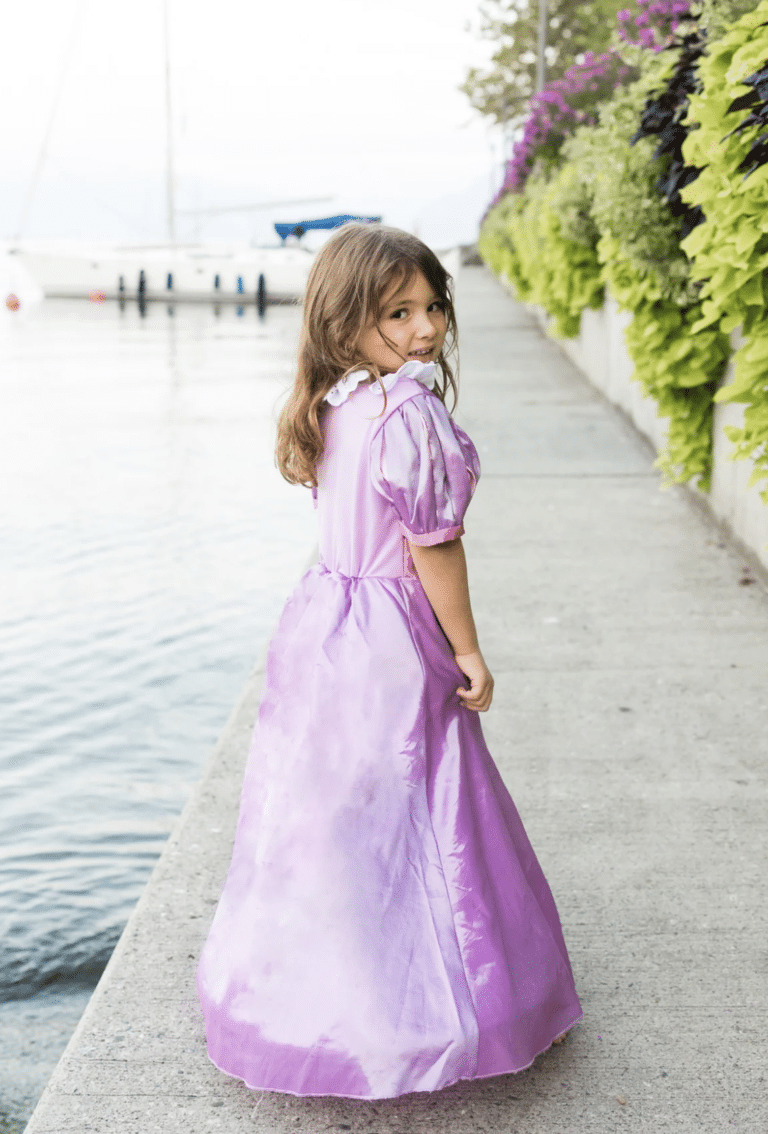 Meisje draagt een paarse Rapunzel verkleedjurk met lange glanzende rok aan het water, van achteren gezien