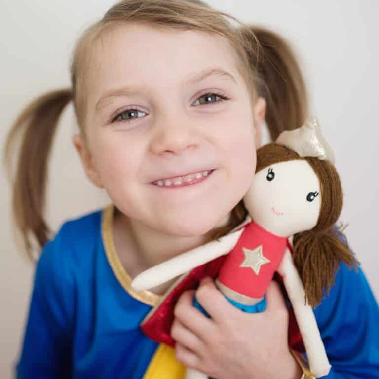 Een meisje houdt de knuffel Suzie the supergirl vast, met rood shirt, blauwe rok, cape en bruin haar.