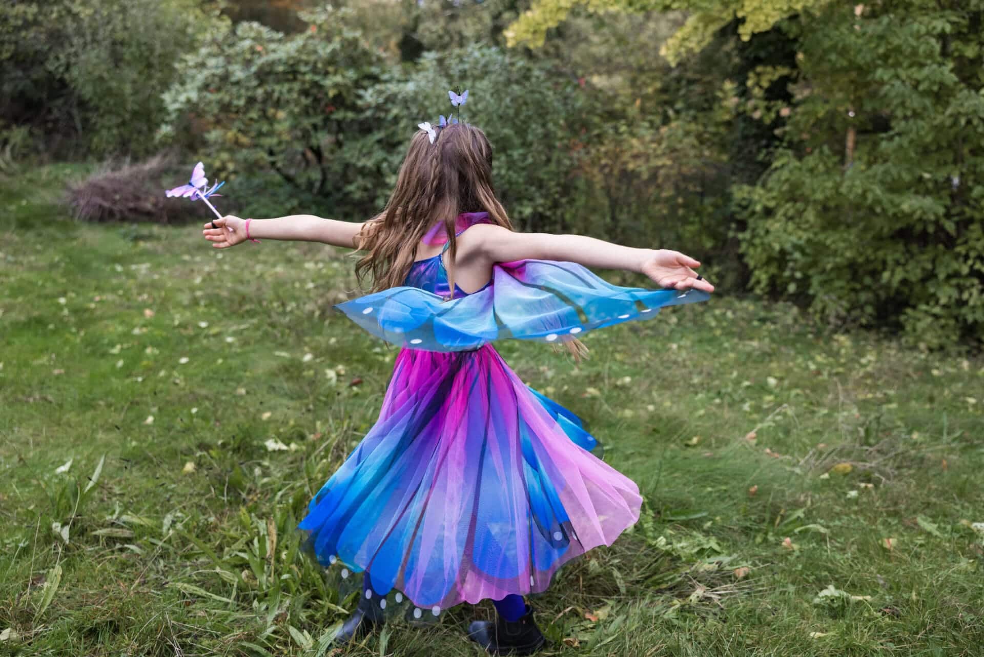 Meisje van achteren in een blauw-paarse verkleedjurk meisje met uitgespreide doorschijnende vlindervleugels in een grasveld.