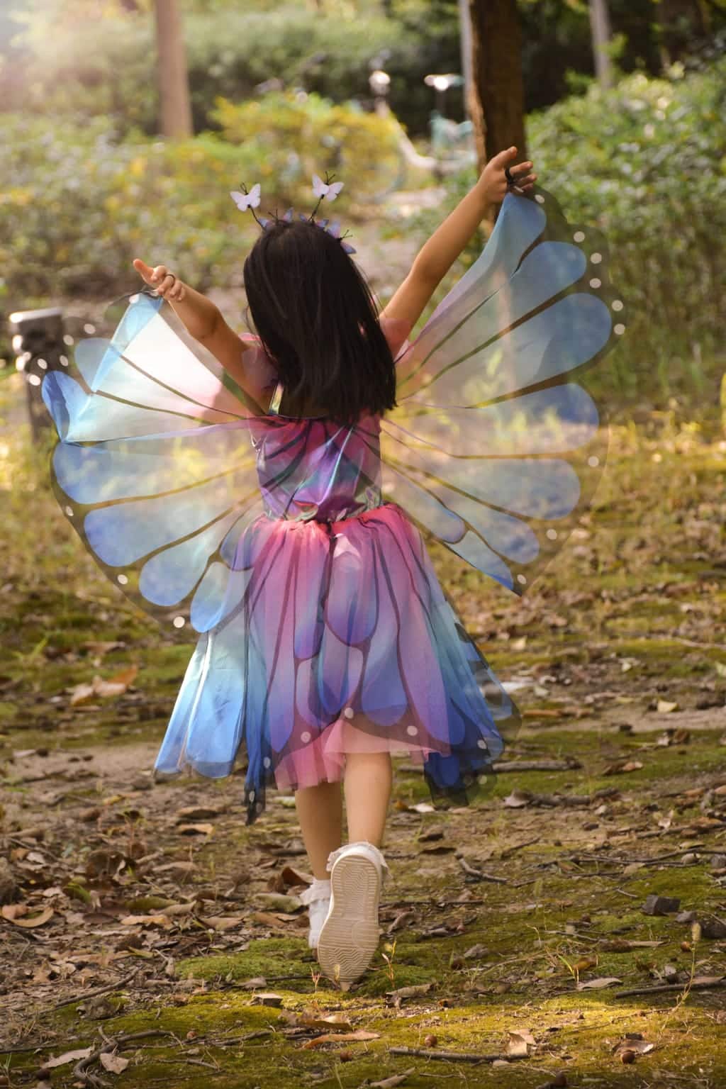 Meisje van achteren in een blauw-paarse vlinderjurk kind met grote doorschijnende vleugels in het bos.
