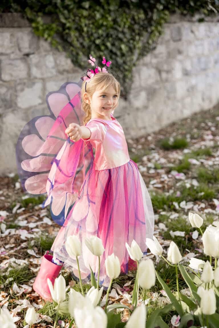 Meisje draagt een roze vlinderjurk met paarse vleugels en bijpassende roze laarzen tussen witte bloemen.