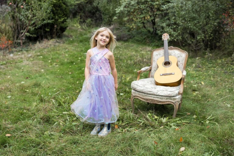 Meisje draagt de lila Ombre Era prinsessenjurk met sterrenversiering in een tuin naast een vintage stoel met gitaar
