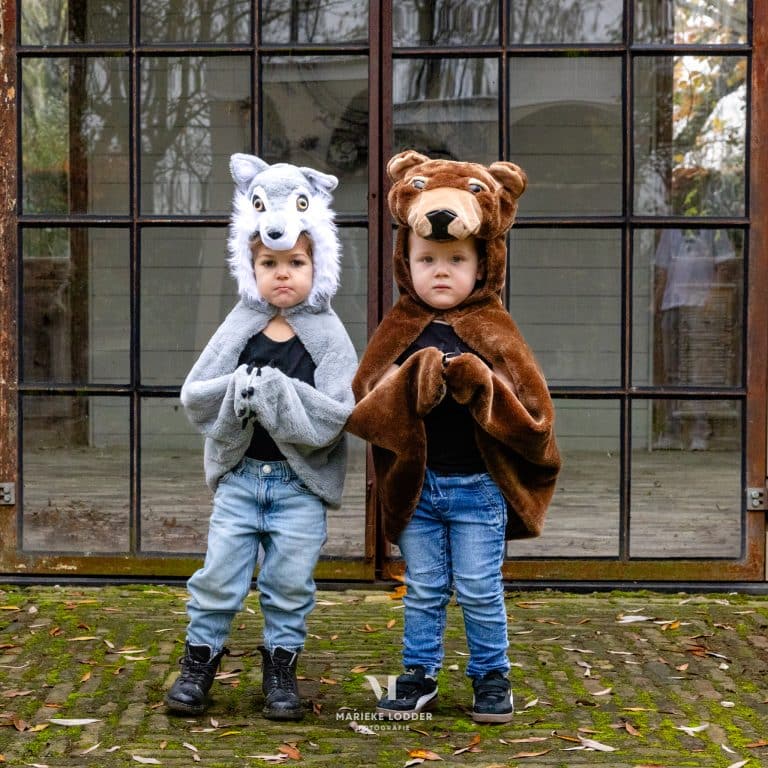 Voor een glazen pui staan twee kinderen frontaal met een grijze wolf cape en bruine bear cape, hun pootjes vooruit.