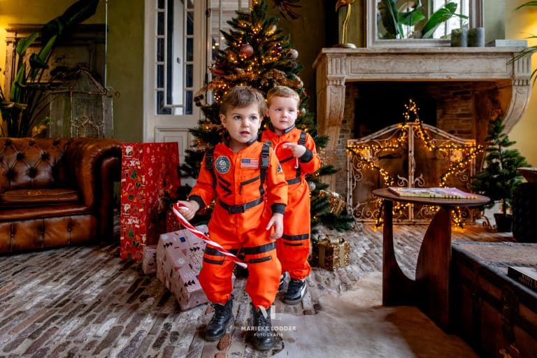 Twee jongens dragen het oranje Astronaut verkleedset bij de kerstboom, met zwarte banden en ruimtepatches.