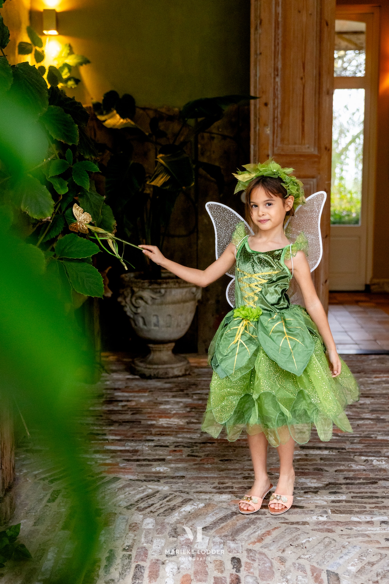 Meisje draagt een groene Tinkerbell jurk met vleugels, bladversiering en glinsterende rok in een sfeervolle hal