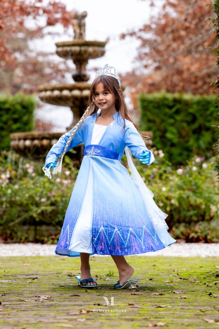 Meisje draagt een blauwe Elsa verkleedjurk met glinsterende rok, witte vlecht en kroon bij een fontein.