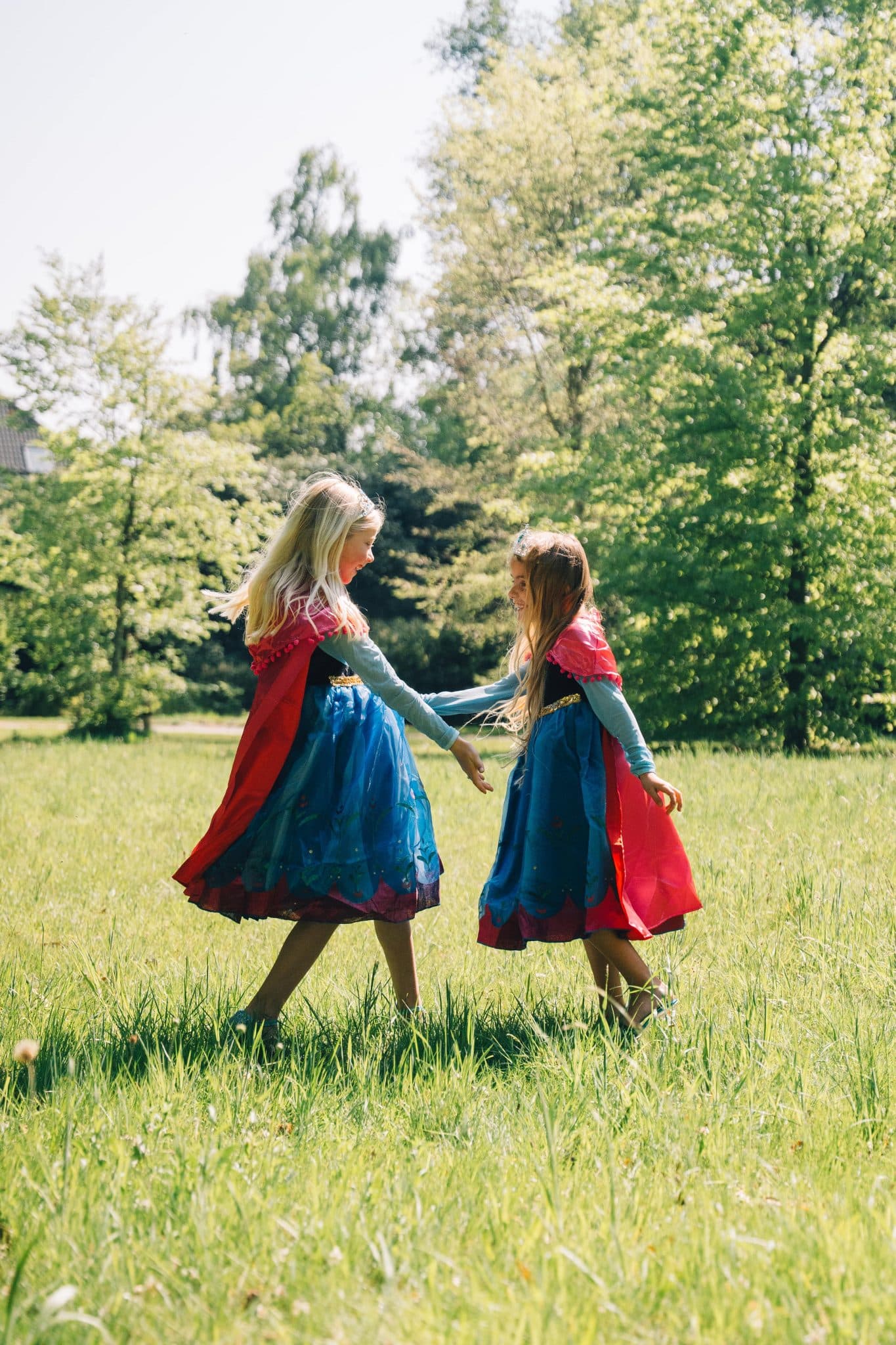 Van opzij lopen twee meisjes in een anna frozen jurk met blauwe rok en rode cape hand in hand door een zonnige weide.