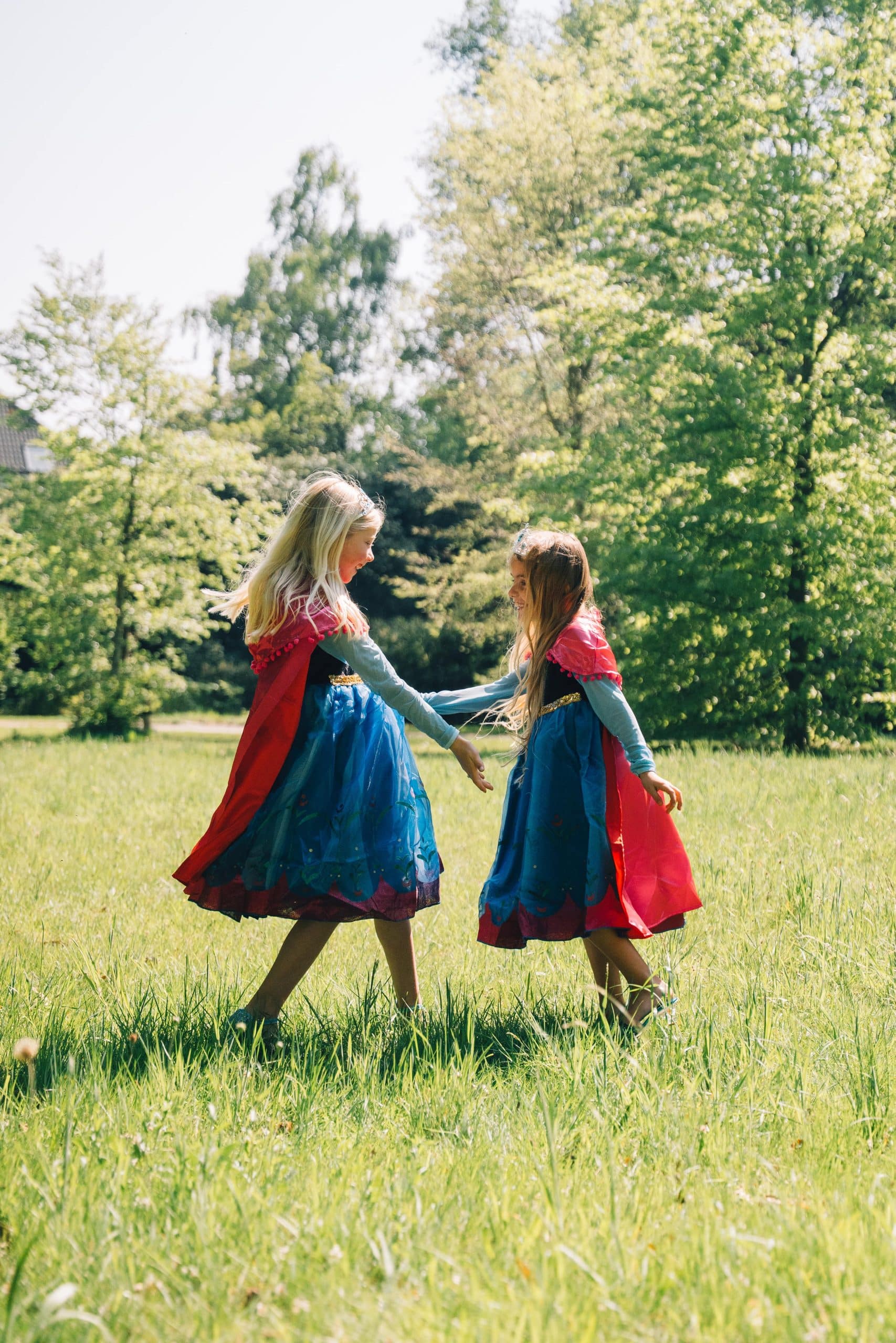 Van opzij lopen twee meisjes in een anna frozen jurk met blauwe rok en rode cape hand in hand door een zonnige weide.