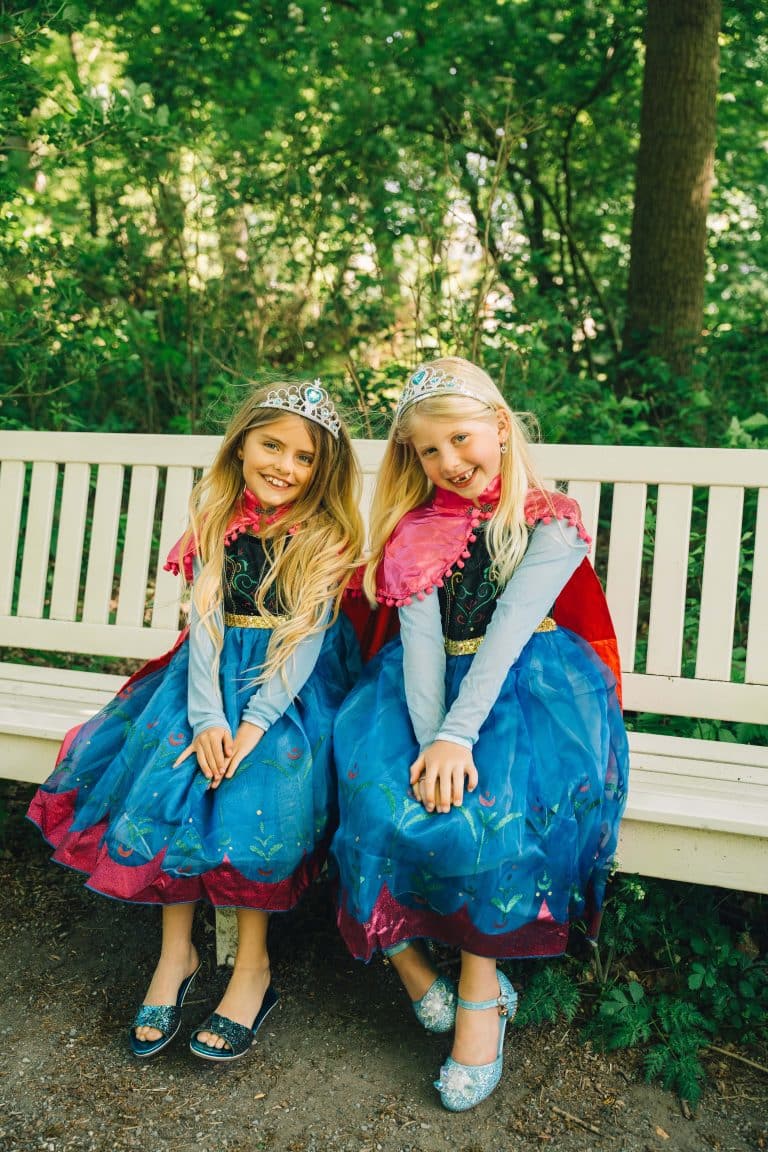 Twee meisjes dragen een anna jurk frozen met blauwe rokken, roze capes en kronen op een witte bank in het bos.