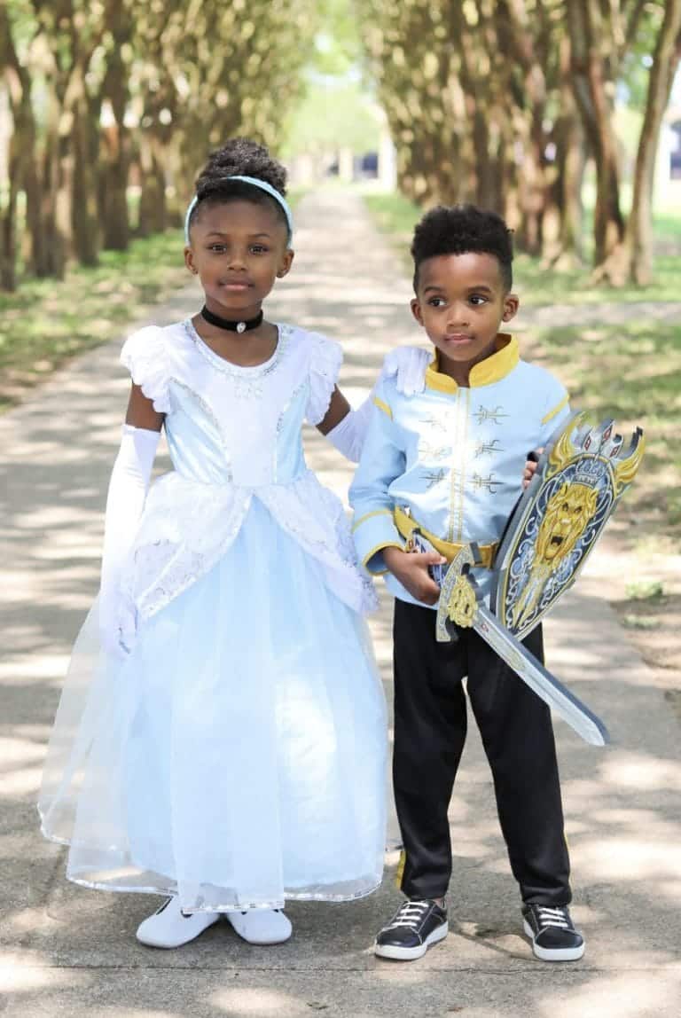 Meisje draagt een lichtblauwe cinderella jurk met witte ruchesmouwen en een lange transparante rok naast een jongen met schild