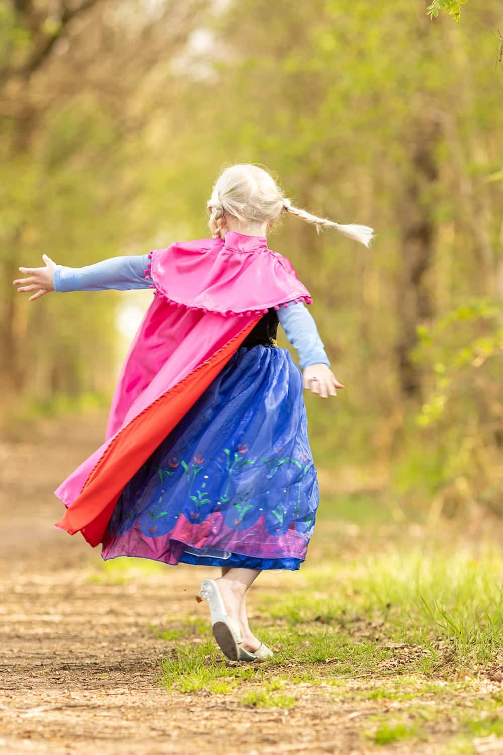 Meisje draagt een Anna jurk met roze cape en blauwe rok terwijl ze over een bospad loopt, van achteren gezien.