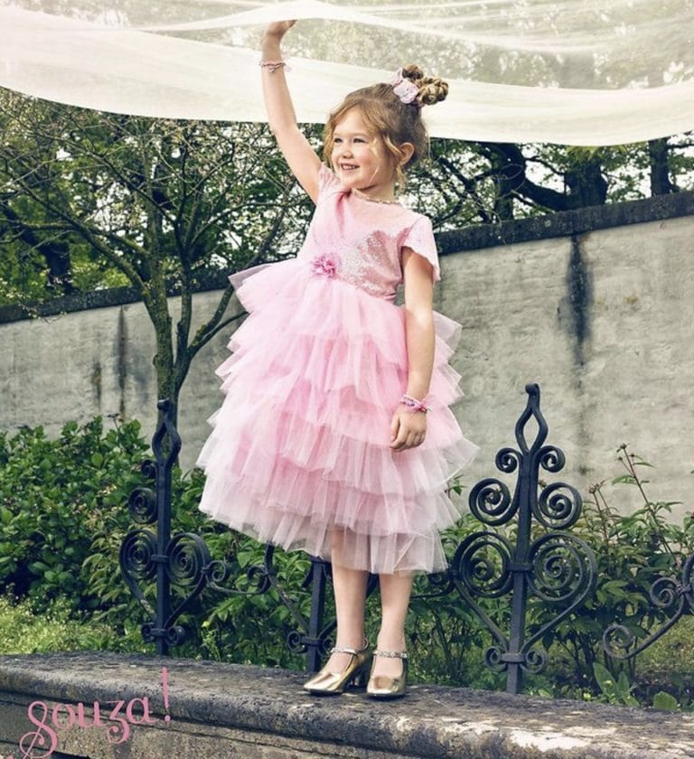 Meisje draagt de roze prinsessenjurk Garance met paillettenlijfje en volle tule rok buiten op een muurtje