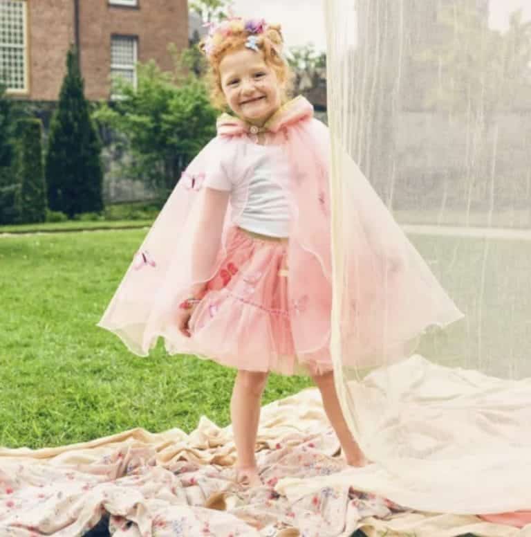 Meisje draagt een roze cape met capuchon en vlinderdecoratie, buiten op een kleed bij een witte tent.
