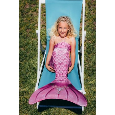 Meisje draagt een roze Kuaki mermaids zeemeerminstaart met glanzende vin, liggend op een blauwe stoel in het gras