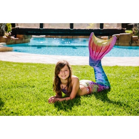 Meisje draagt een Kuaki zeemeerminstaart met regenboogkleuren en bikinitop, liggend op het gras bij een zwembad.