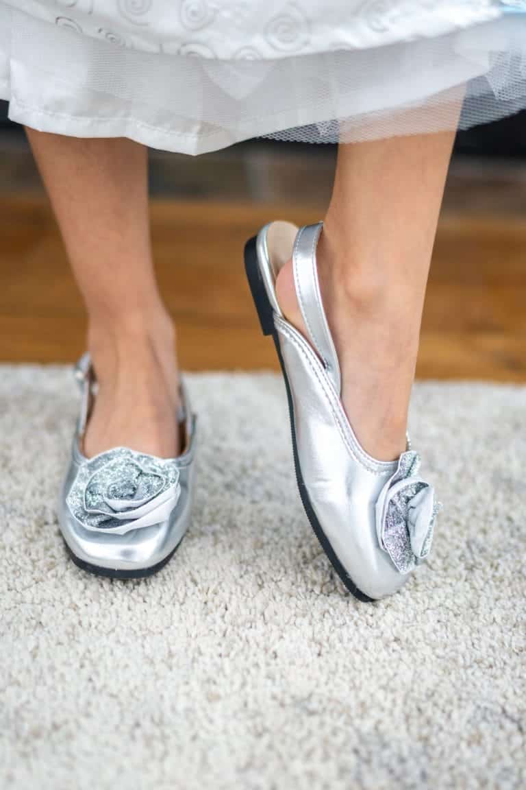 Zilveren kinderschoenen aan de voeten van een kind, met glitterrozetten, bandjes en zwarte zolen onder een jurk