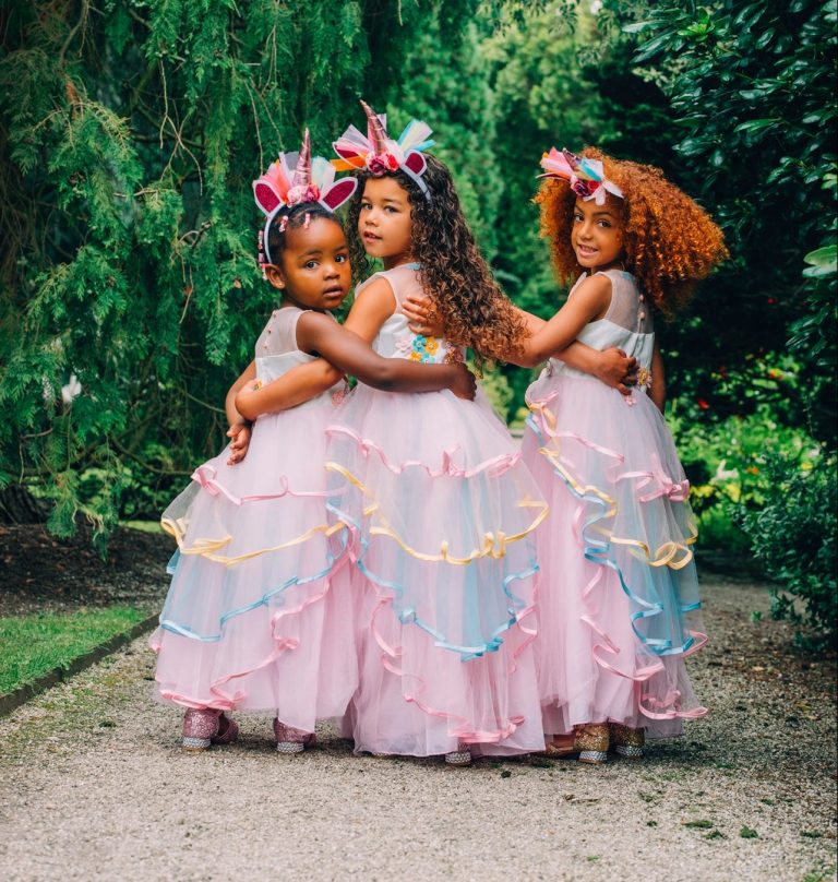 Drie meisjes staan omarmd in roze unicorn jurken met kleurrijke linten en eenhoornhaarbanden, gezien van achteren in een tuin.