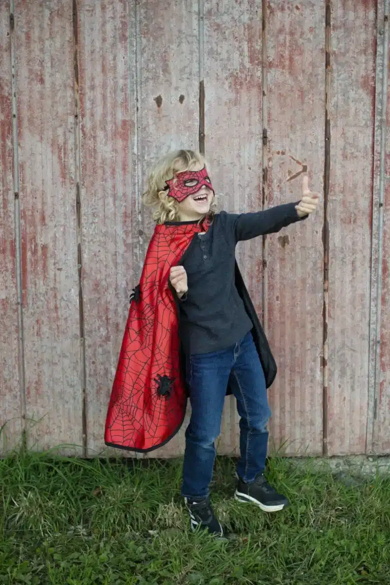 Kind draagt een rood superhelden kostuum met masker en cape met spinnenwebprint en geeft een duim omhoog