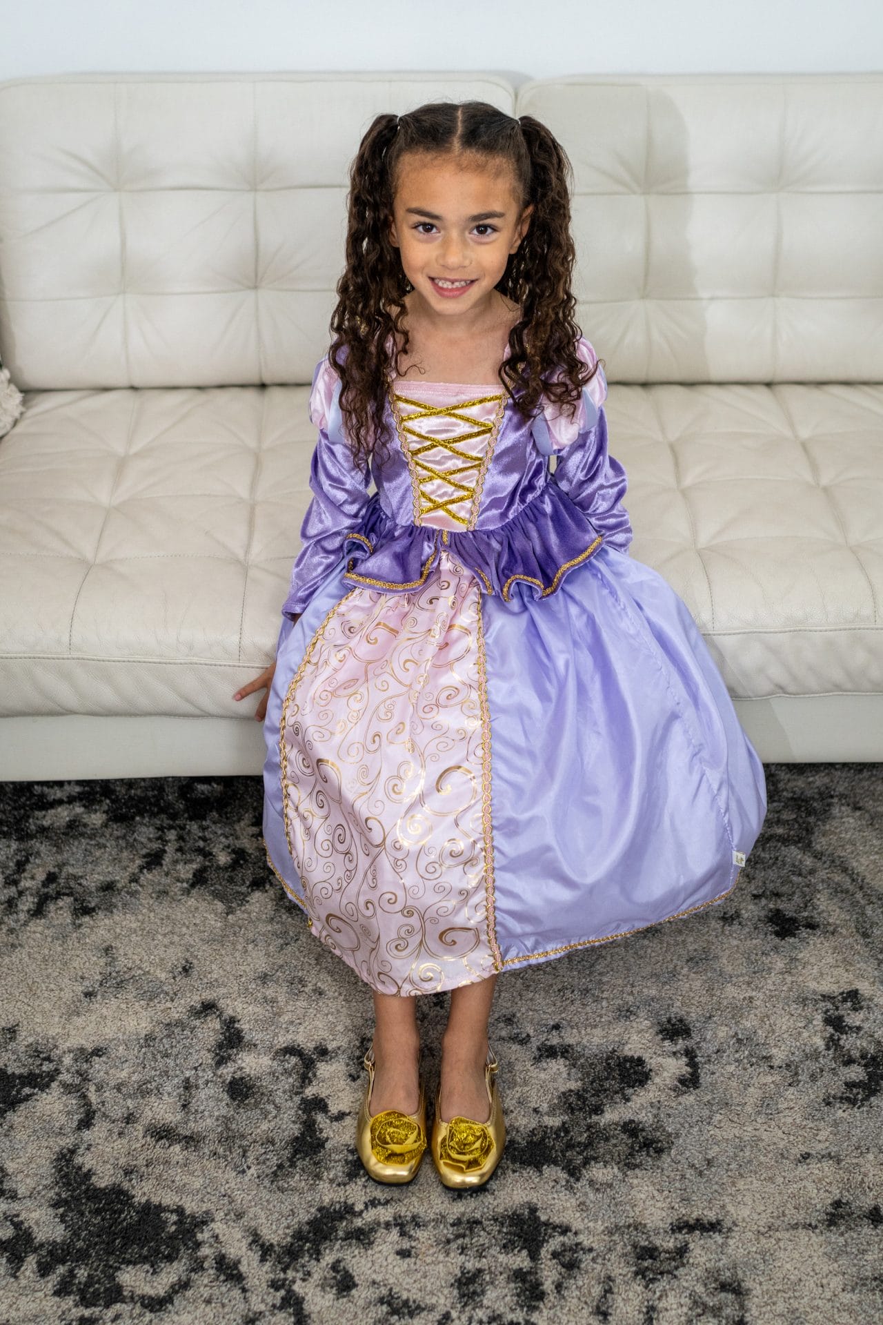 Een meisje draagt gouden kinderschoenen van Little Adventures met glanzende rozetten onder een paarse prinsessenjurk.