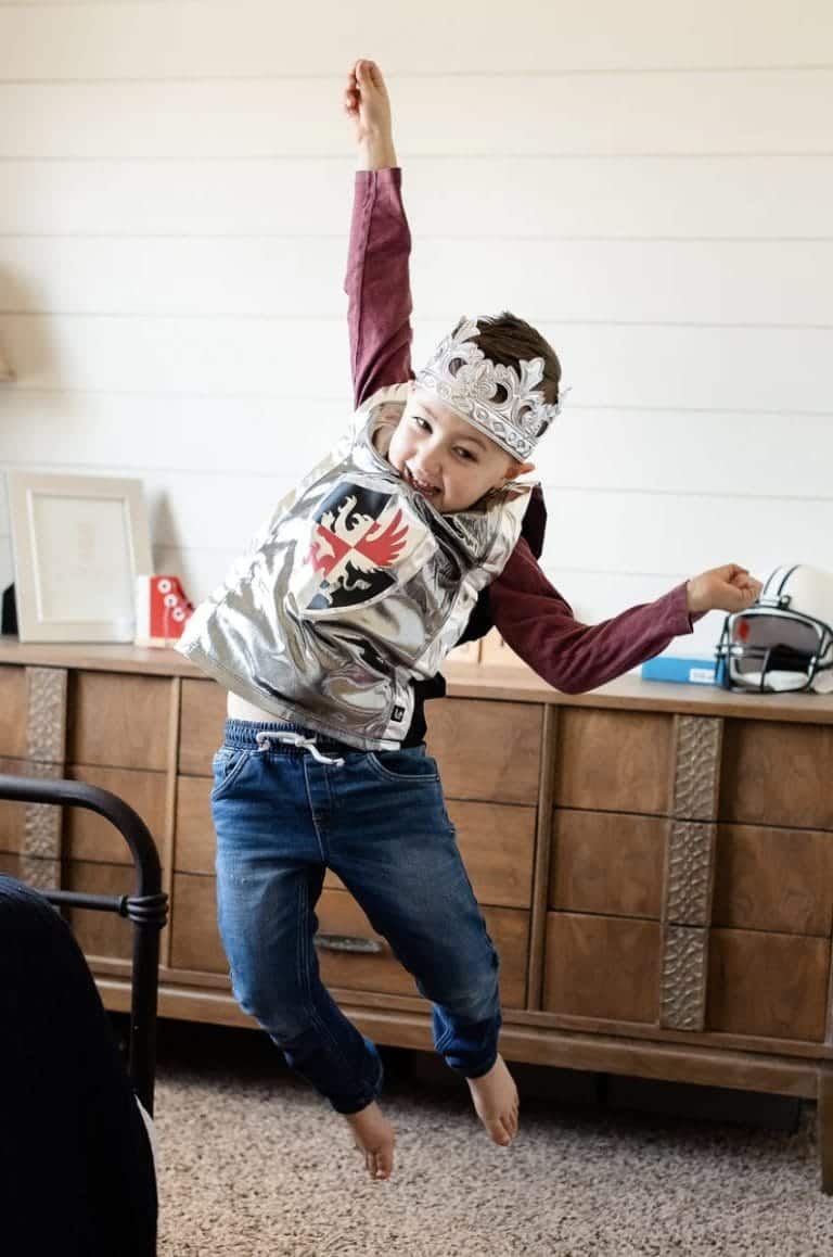 Jongen draagt een glanzend zilveren ridder vest met zwart-rood schild en maakt een stoere pose.