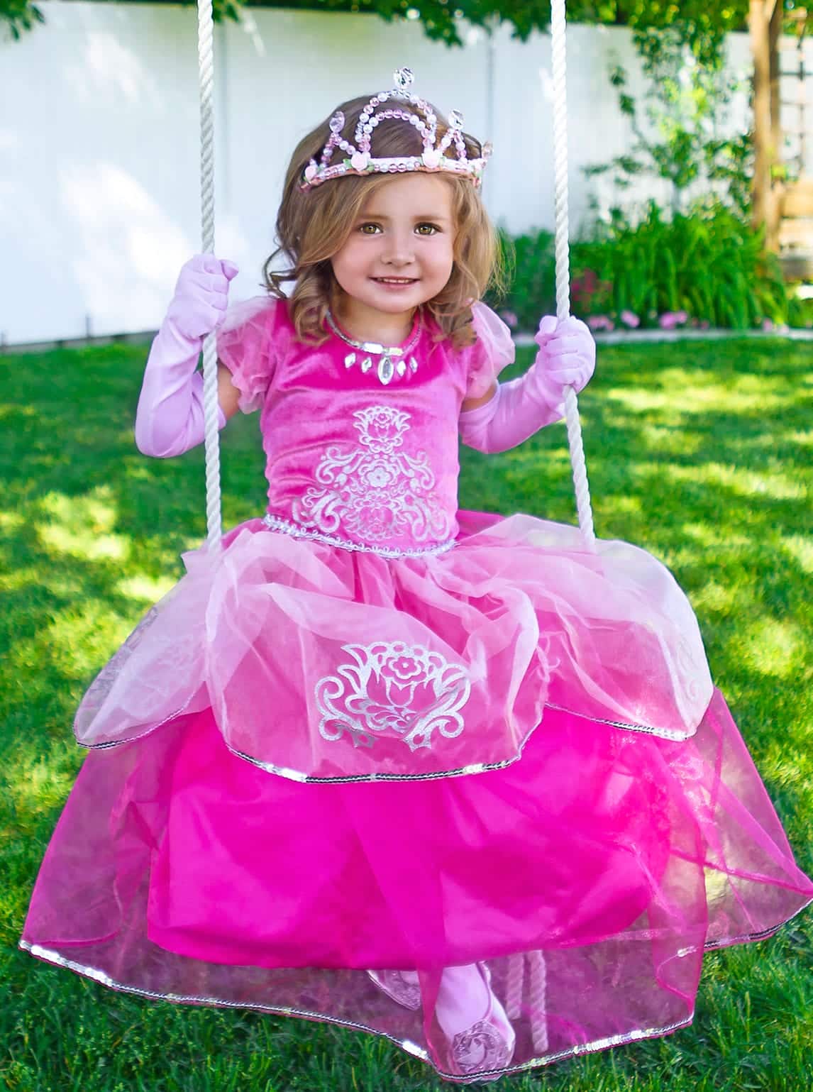 Meisje op een schommel draagt de fuchsia Deluxe Pink prinsessenjurk met zilveren versieringen en roze kroon.