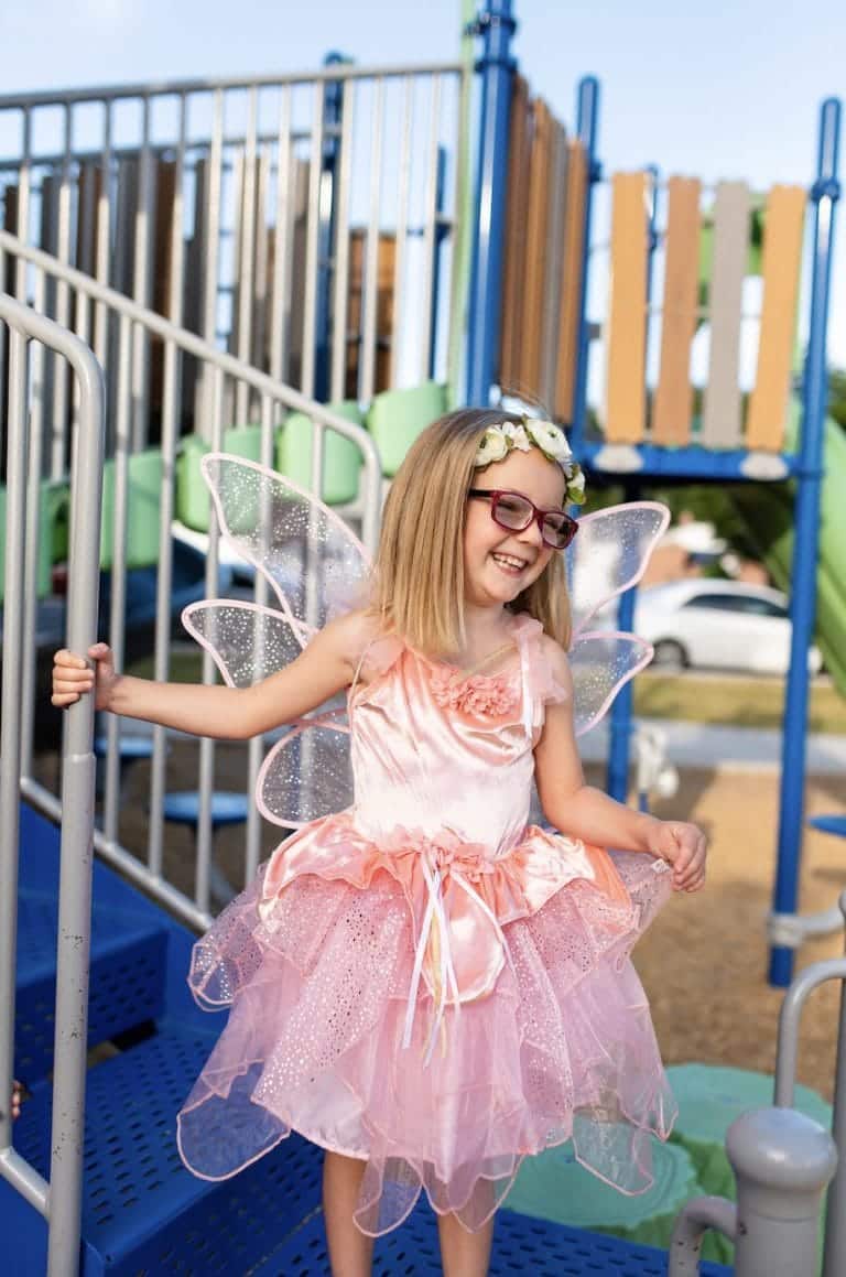 Meisje draagt een roze feeen jurk van Little Adventures met bloemen, tule rok en vleugels op een speelplein.