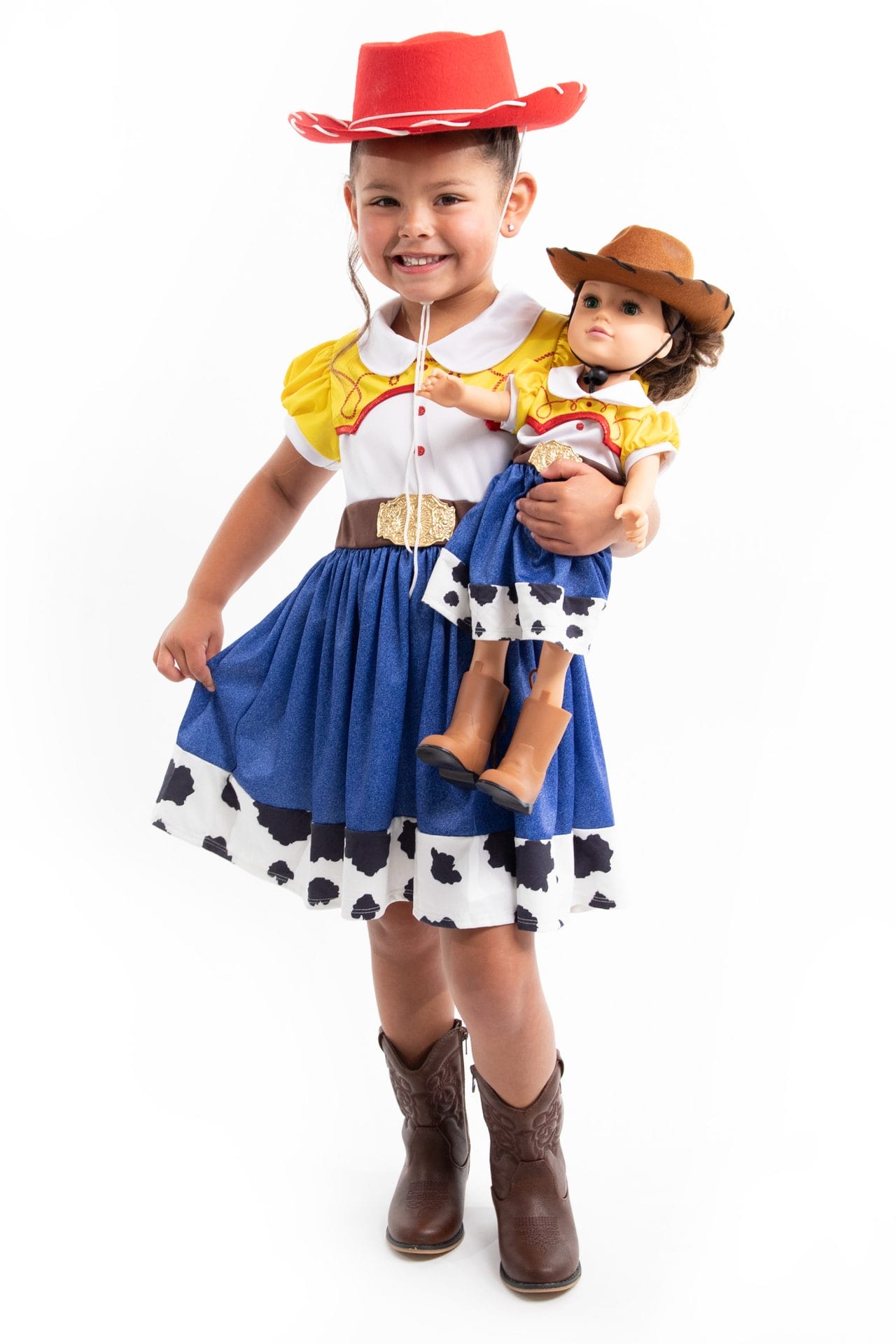 Vooraanzicht van een meisje in een Toy Story-cowgirljurk met koeienprint, dat een pop vasthoudt tegen een witte achtergrond.