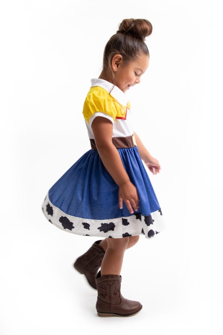 Meisje draagt een Toy Story-cowgirljurk met geel-wit lijfje, bruine riem, blauwe rok met koeienprint en cowboylaarzen.