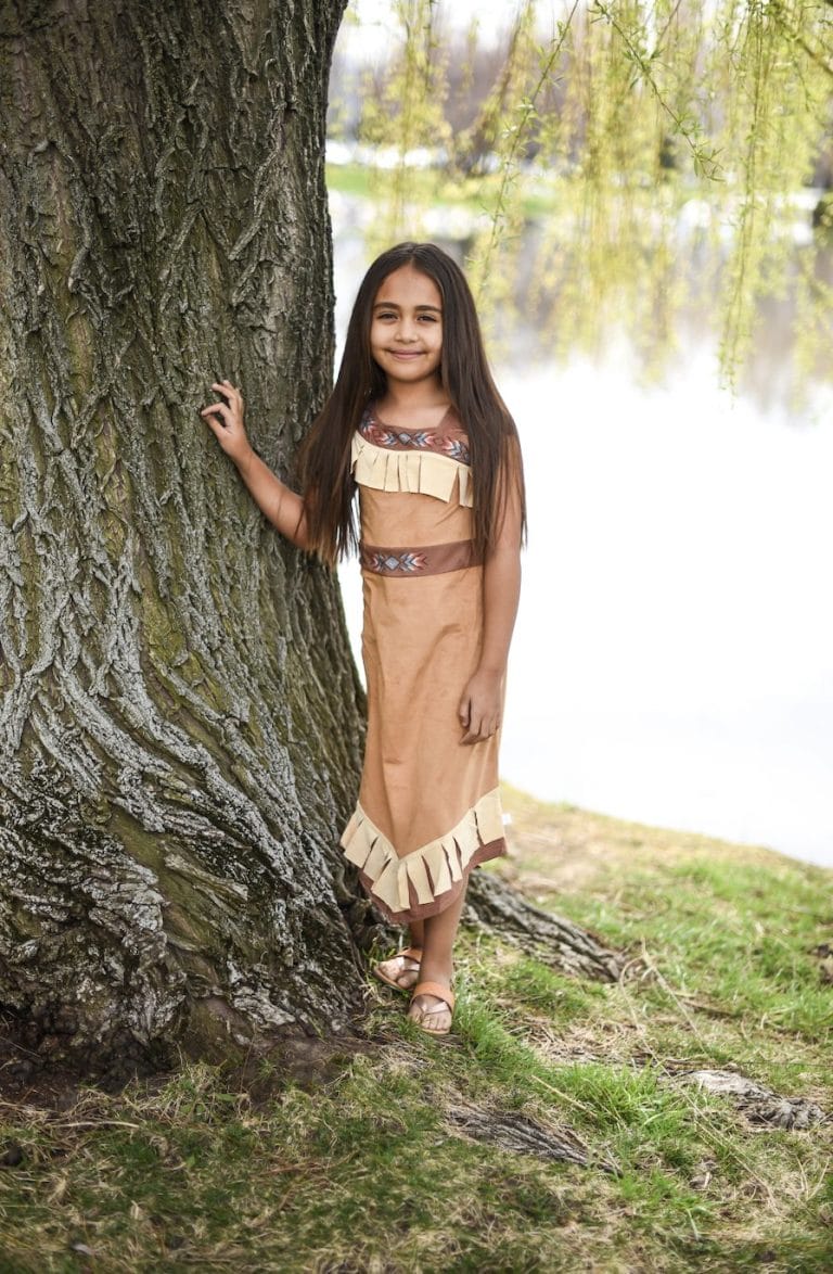 Meisje draagt de bruine Woodland Princess princessenjurk van Little Adventures naast een boom, met franjes aan de zoom.
