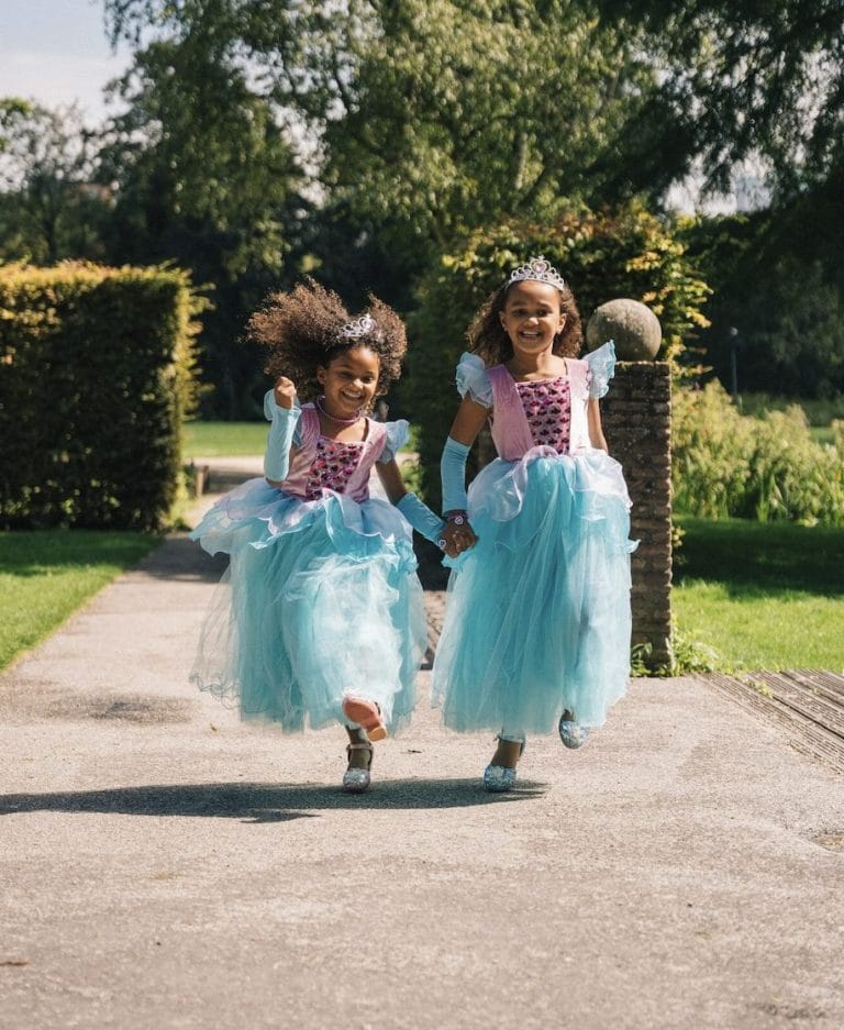 Twee meisjes dragen blauwe zeemeermin jurken met tule rokken, handschoenen en kroontjes in een zonnige tuin.