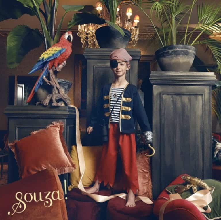 Jongen draagt het Souza! piratenpak met blauw jasje, rode broek, rode bandana en zwart ooglapje in een sfeervol interieur.