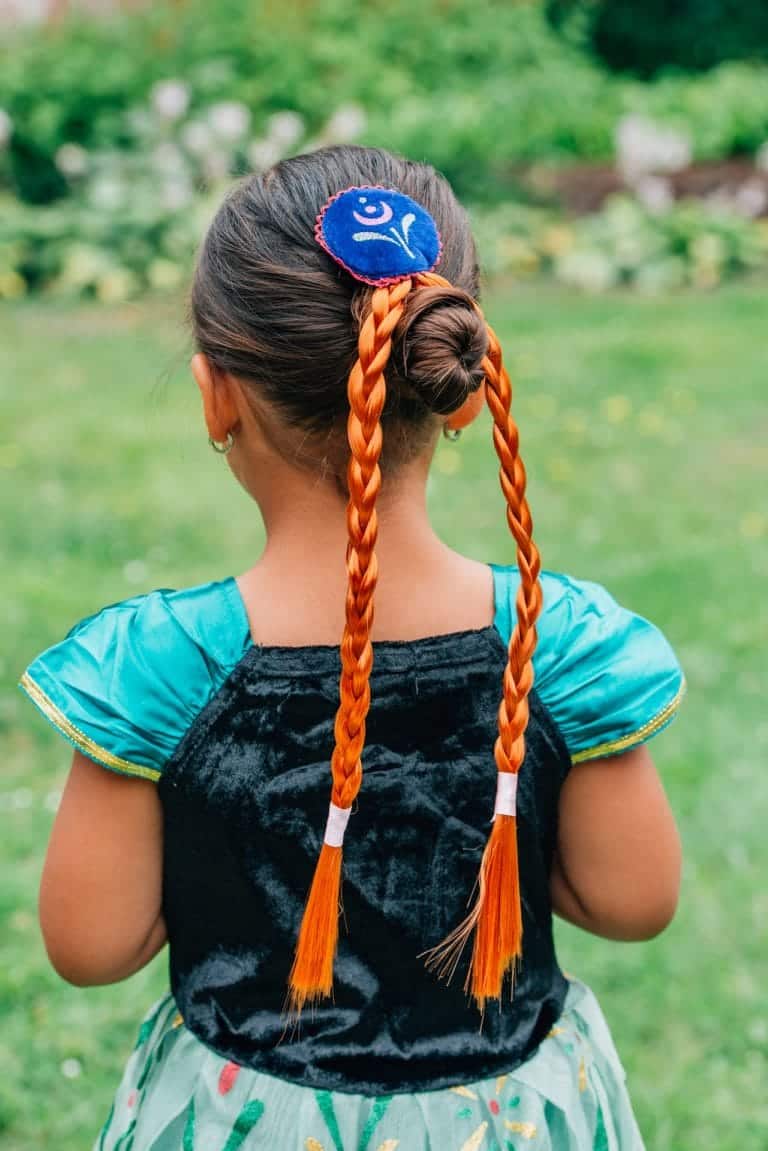 Meisje draagt de bruine Anna vlecht met haarclip en Frozen-broche bij een blauwe prinsessenjurk, van achteren gezien