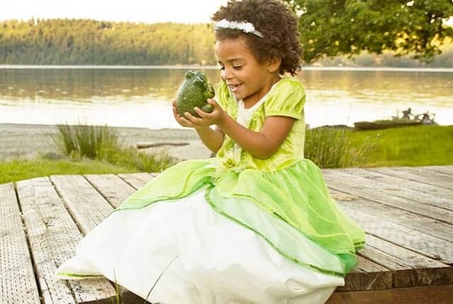 Meisje draagt een groene prinses Tiana jurk en zit op een houten steiger bij het water met een groene kikker in haar handen.