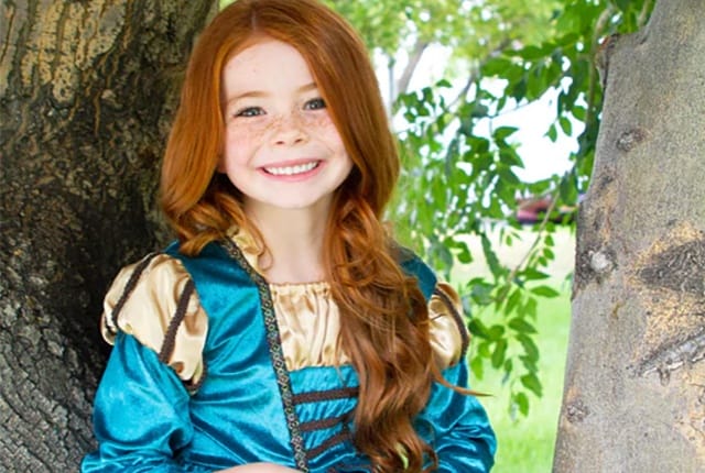 Een meisje draagt een blauwe Merida jurk met gouden mouwen en bruine details, tussen de bomen buiten.