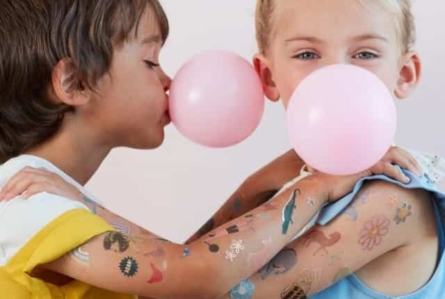 Twee kinderen tonen kleurrijke kindertattoos op hun armen terwijl ze roze ballonnen opblazen.