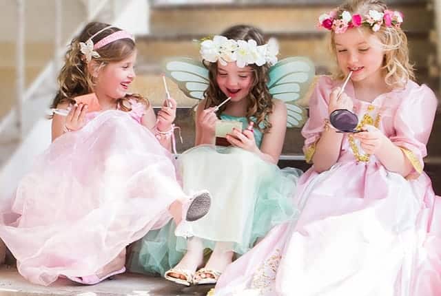 Drie meisjes gebruiken kindermake-up in roze en mintgroene prinsessenjurken, met bloemenkransen en vleugels.