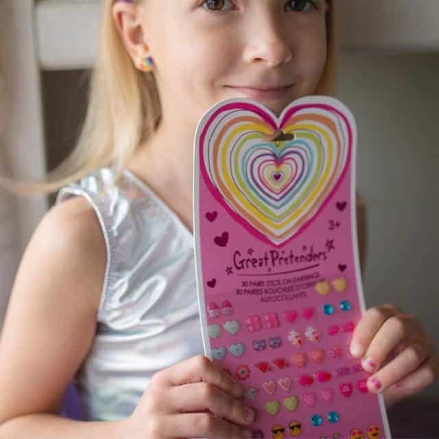 Meisje toont een roze kaart met 30 paar Rainbow Heart plak oorbellen in kleurrijke hartvormen.