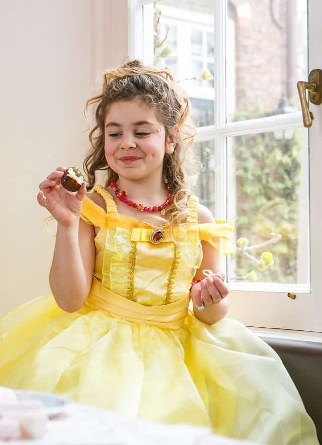 Meisje draagt de gele prinsessenjurk Belle van Het Betere Merk en houdt een koekje vast bij het raam.
