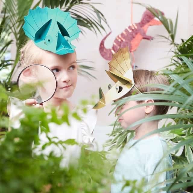 Twee kinderen dragen elk een dino verjaardagsmuts van Meri Meri tussen tropische planten, met een vergrootglas in beeld