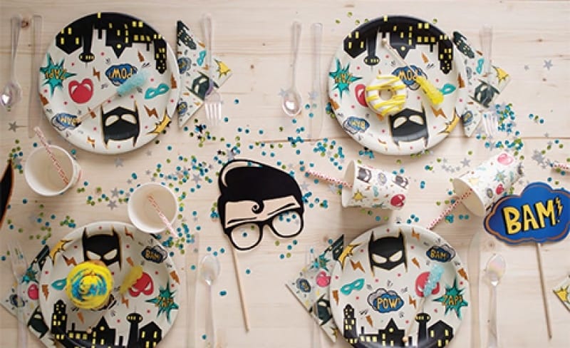 Superhero servetten op een feesttafel met superheldenborden, maskers, confetti en een grote BANG!-tekst.