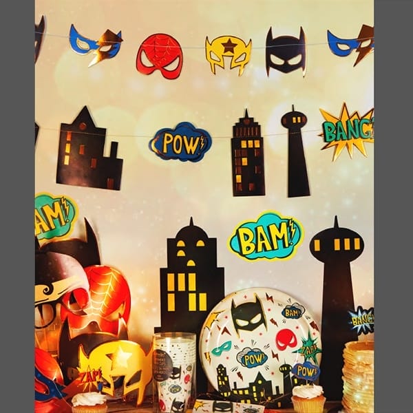 Superhero feestslinger Gebouwen met zwarte silhouetten en POW!- en BANG!-teksten boven een feesttafel.