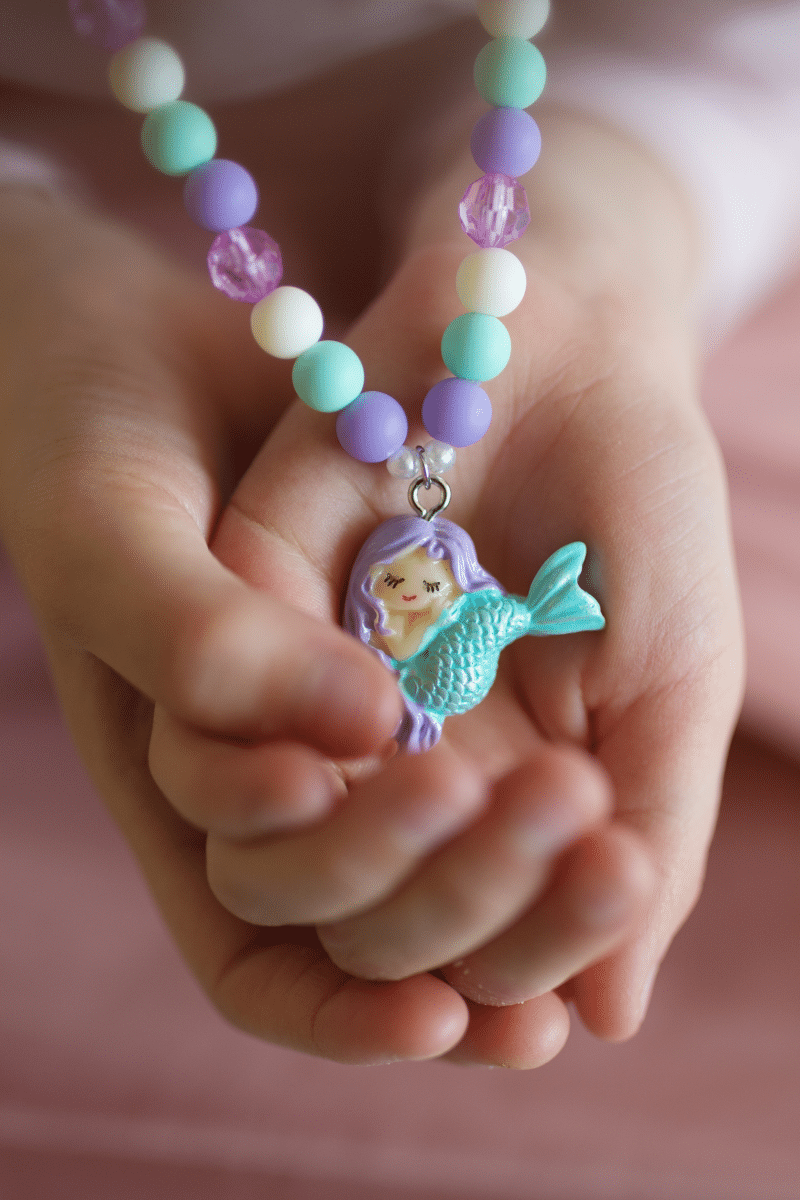 Close-up van een kinderhand die een zeemeerminketting vasthoudt, met pastelmatte kralen en een turquoise zeemeerminbedel.