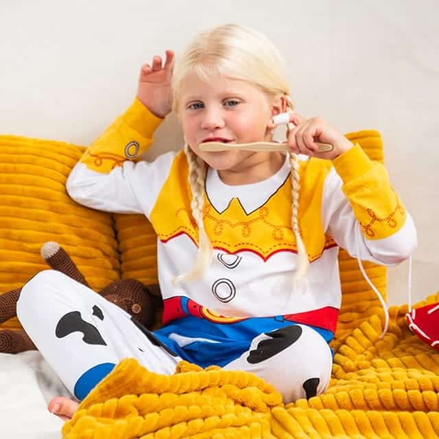 Meisje draagt een geel-witte Toy Story pyjama met cowboydetails, zittend op een gele bank.