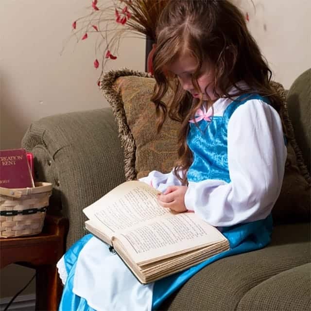 Meisje leest in een boek terwijl ze een blauwe Belle jurk met witte mouwen en roze borststrik draagt.